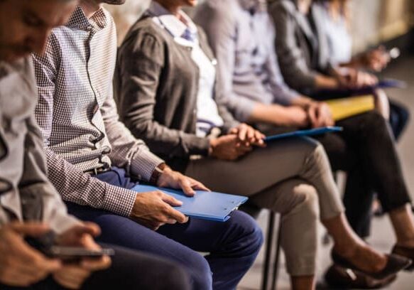 Large group of unrecognizable business people waiting for a job interview. Focus is on man with blue clipboard.